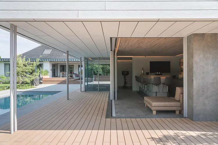 Moderne offene Terrasse eines Poolhauses mit Holzdeck, die nahtlos in einen Innenraum mit Bar und Sitzgelegenheiten übergeht. Im Hintergrund sieht man einen Pool und ein Wohnhaus mit großen Glasflächen, umgeben von einer gepflegten Grünanlage.