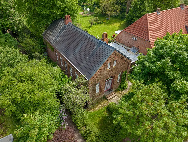 Professionelle Drohnenaufnahme eines Backsteinhauses mit dunklem Schieferdach, umgeben von üppigem Grün und einem gepflegten Garten. Die Vogelperspektive zeigt die Architektur und die idyllische Umgebung – ideal für Immobilienpräsentationen oder Landschaftsdokumentationen.