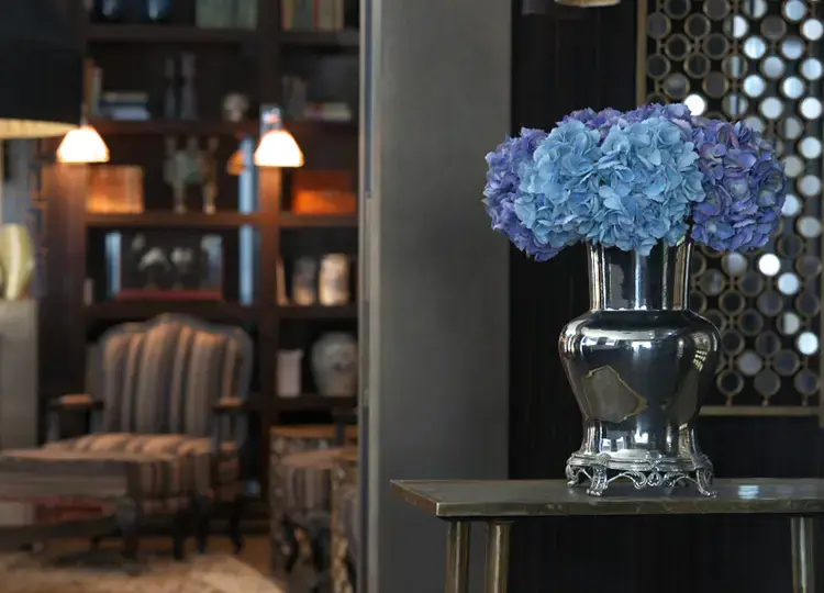 Eine elegante Vase mit blauen Hortensien auf einem schlichten Tisch, umgeben von einer gemütlichen Leseecke mit Sessel, Bücherregal und warmem Licht – perfekt für hochwertige Präsentationen von Wohnräumen oder Hotelinterieurs.