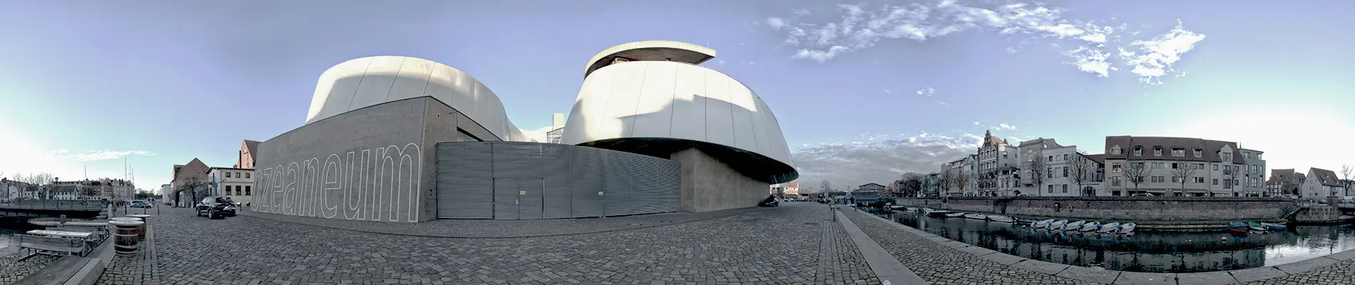 Hochwertige 360°-Fotografie für einen virtuellen Rundgang: Eine beeindruckende Panoramaaufnahme eines futuristischen Gebäudes mit organisch geschwungenen, glatten Oberflächen aus hellem Material. Die Architektur besticht durch klare Linien und eine dynamische Formensprache. Im Vordergrund ein Kopfsteinpflasterweg, der den Kontrast zwischen Moderne und Tradition unterstreicht. Im Hintergrund historische Gebäude und ein ruhiger Wasserlauf, die die harmonische Verbindung von Alt und Neu zeigen. Die präzise Ausführung der 360°-Aufnahme ermöglicht eine immersive Erfahrung.